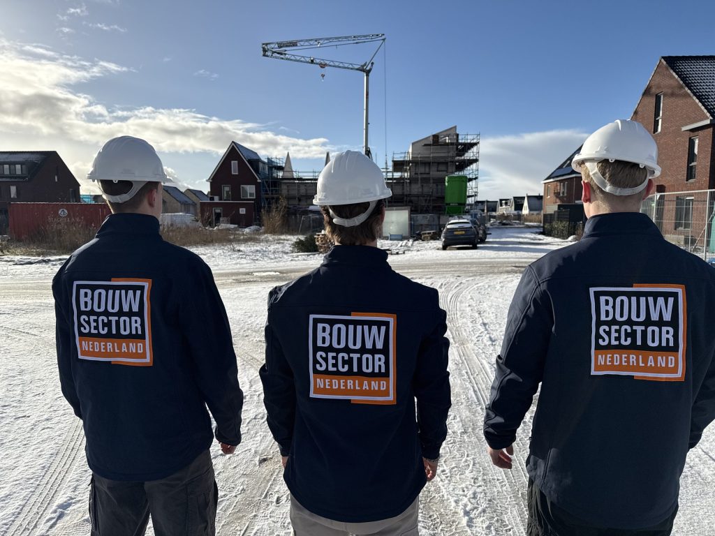 Drie collega's in werkkleding met oranje-wit logo overzien samen een bouwplaats